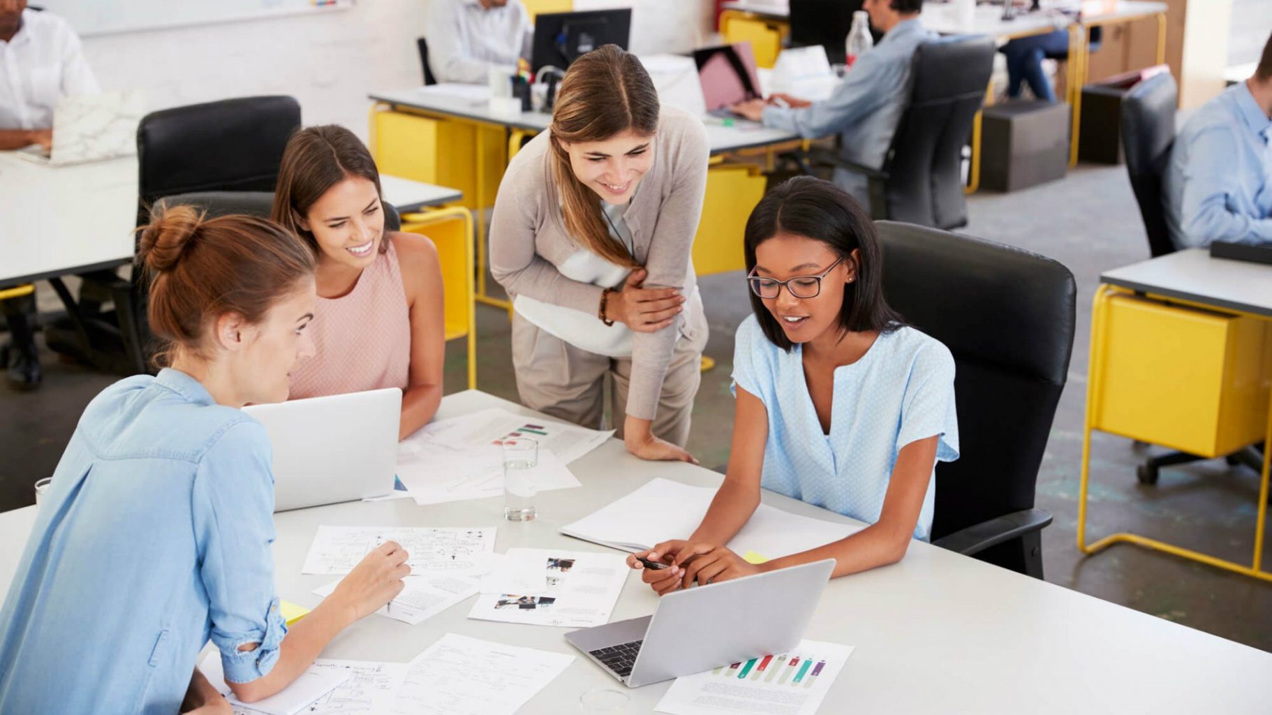 Quatro mulheres conversando ao redor de uma mesa e de um notebook, vestidas socialmente para simular uma avaliação 360