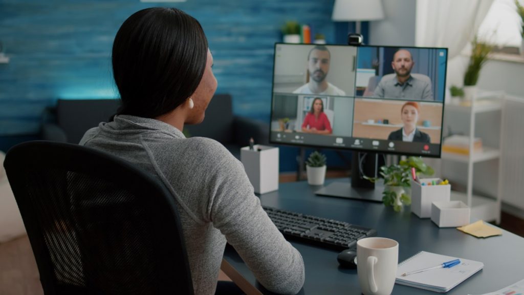 Mulher negra com blusa cinza em uma mesa fazendo uma vídeo conferência simulando uma gestão de equipes híbridas