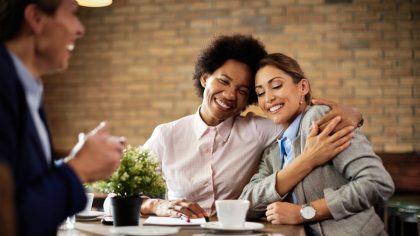 mulheres com roupas sociais, uma de longe e outras duas se abraçando e rindo em volta de uma mesa para representar a empatia no ambiente de trabalho