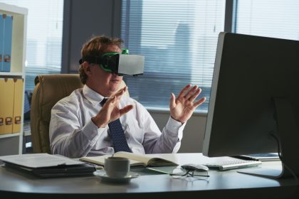 Homem sentado à mesa de escritório, com um computador a frente e utilizando óculos de realidade virtual para simbolizar a gamificação