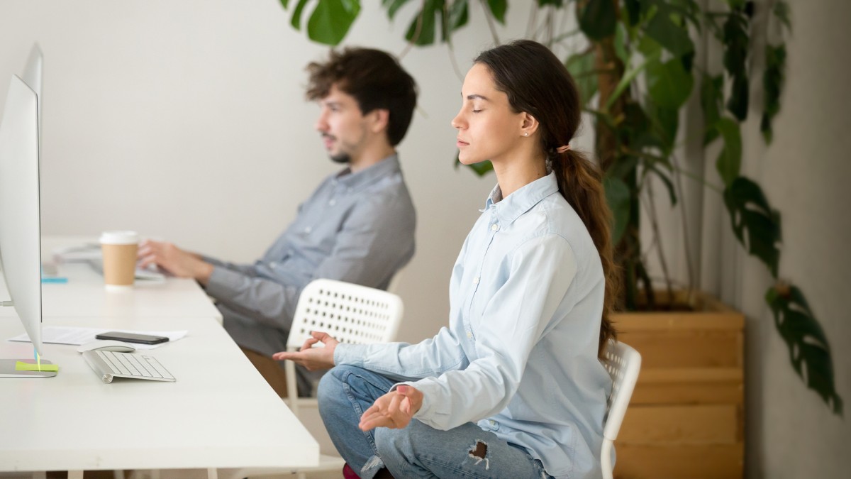 Um homem e uma mulher sentados, meditando, em frente à uma mesa com computadores para simbolizar a saúde mental no ambiente de trabalho