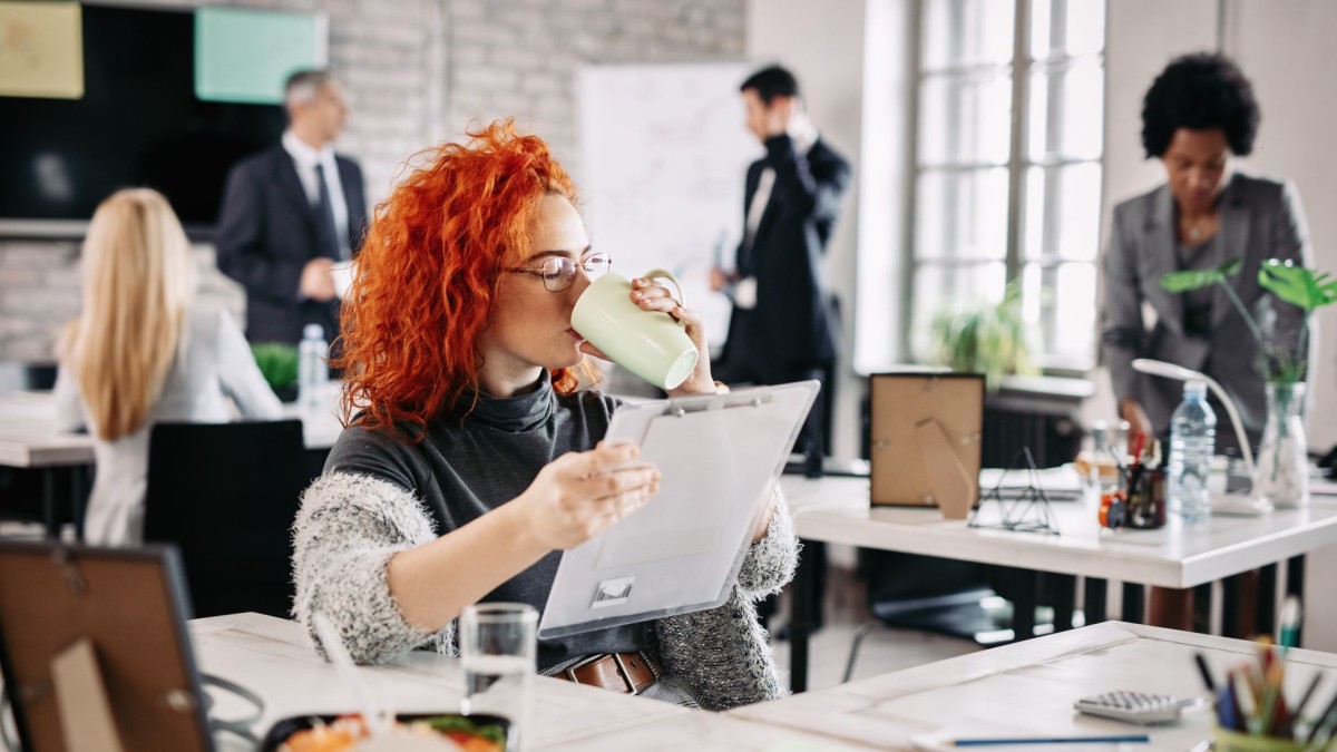 foto de uma sala com profissionais em pé e sentados e à frente uma mulher ruiva tomando café e olhando uma prancheta para representar a autonomia no trabalho