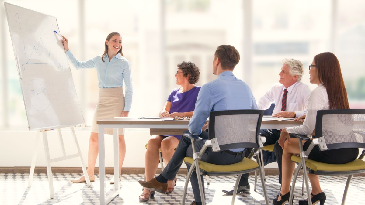 foto de executivos ao redor de uma mesa, assistindo uma mulher apontando para um quadro branco para simbolizar LNT: levantamento das necessidades de treinamento