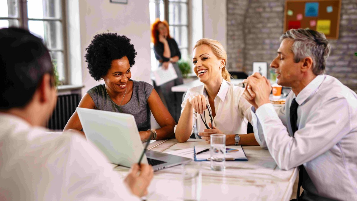 Pessoas com roupas executivas, em volta de uma mesa, conversando e sorrindo para representar uma reunião de trabalho de forma descontraída e a qualidade de Vida no Trabalho