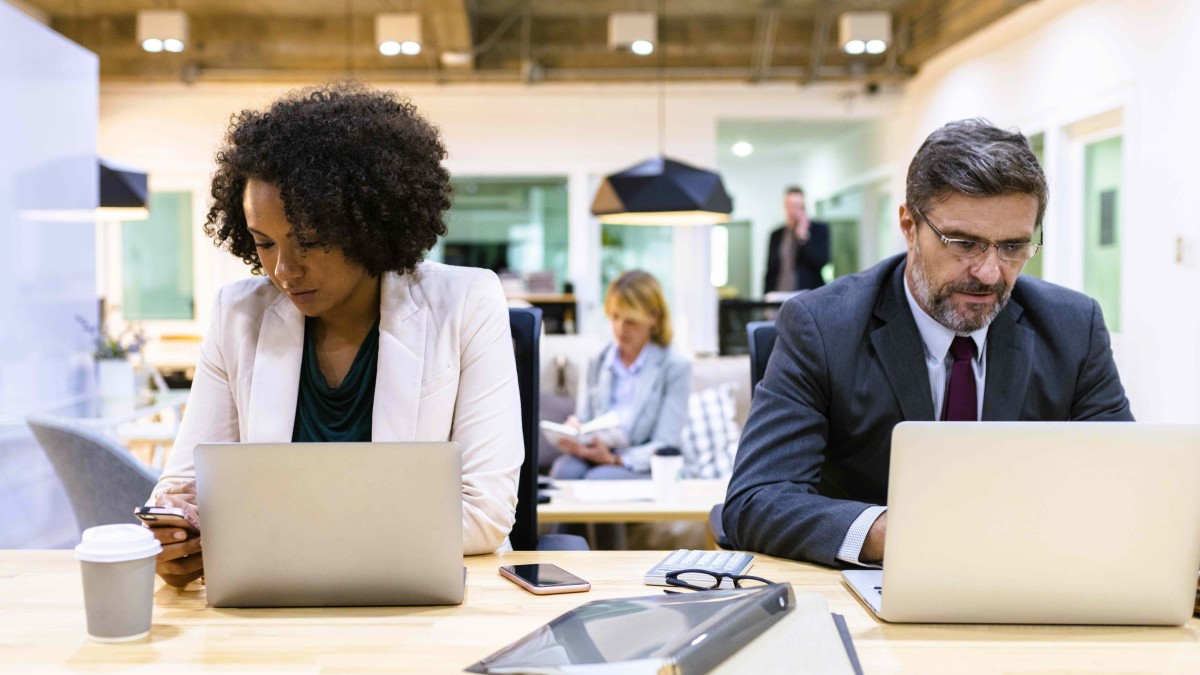 Uma mulher negra e um homem, ambos com roupa social, usando notebook, trabalhando em uma bancada de um espaço de trabalho compartilhado