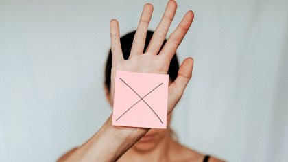 Imagem de uma mulher desfocada; em primeiro plano, a mão aberta mostra um papel com um X