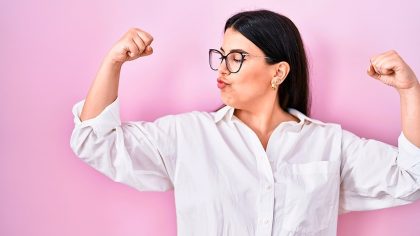 Em fundo rosa, mulher branca de cabelo pretos usando camisa social branca levanta os braços contraindo ambos bíceps, mostrando o muque