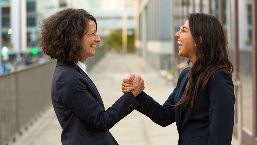 Duas mulheres brancas com roupas corporativas em uma ambiente externo segurando a mão uma da outra demosntrando confiança e parceria