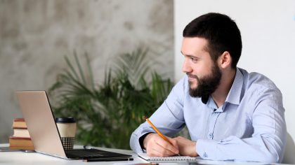 Homem branco em ambiente corporativo, sentado à mesa de frente para o notebook, vendo a tela e fazendo anotações em um bloco de papel