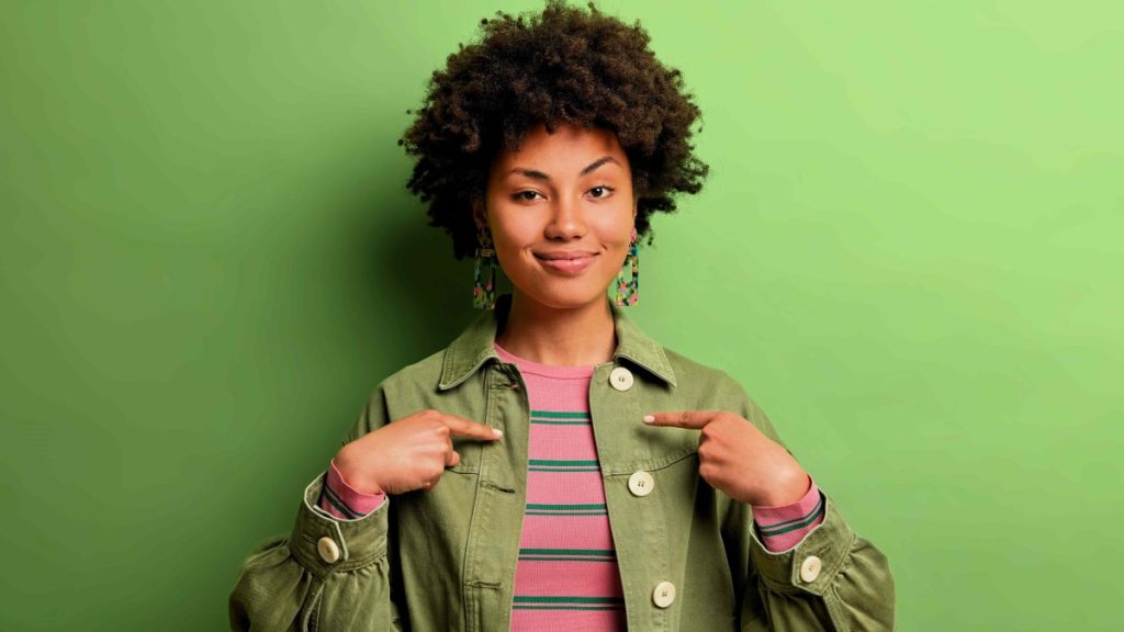 Em fundo verde, mulher negra posicionada em plano americando apontando para si mesma, demonstrando protagonismo