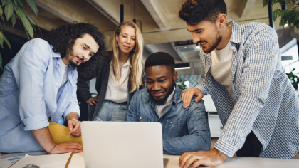 Quatro colegas de equipe trabalham juntos em um projeto, concentrados ao redor de um laptop em um ambiente de escritório moderno. A expressão de engajamento de todos destaca a colaboração e a importância do trabalho em equipe para alcançar objetivos comuns.