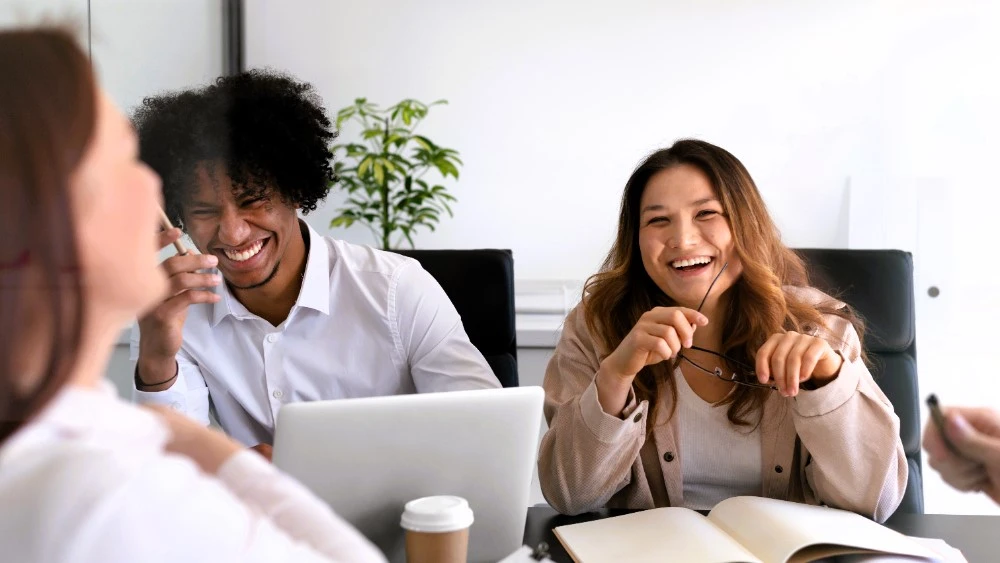 Grupo de colegas em um ambiente de trabalho, rindo e interagindo de forma descontraída durante uma reunião, transmitindo um clima de colaboração e bem-estar