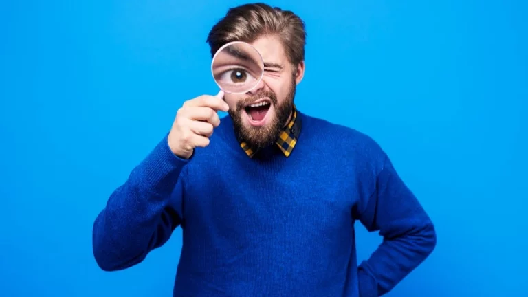 homem caucasiano vestido com casaco azul em um fundo azul, segurando uma lupa na mão, o que deixa seu olho ampliado.