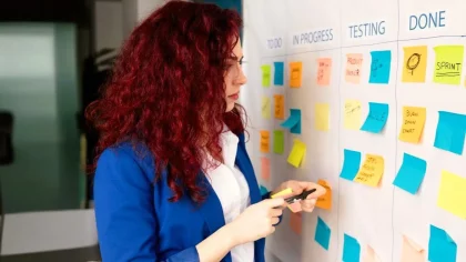 Mulher em ambiente corporativo em frente a um quadro branco cheio de papéis adesivos coloridos, distribuidos em colunas diferentes, indicando atenção ao planejamento pessoal.